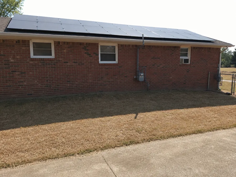 The shadow of a roofline and its row of solar modules thrown across dry grass, with the brick house and its full module row behind