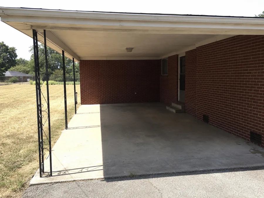 The open carport of a single storey red brick ranch house, hard summer sun across the concrete apron meeting a sharp line of shade beneath the roof